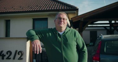 Evin önündeki konut kapısına yaslanan adam, gülümseyen ve rahat, banliyö yaşam tarzı ve mülk çevresi odaklı