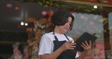 Tabletteki dijital içeriği inceleyen çiçekçi, çiçekçi dükkanının önünde dikiliyor, çiçeklerle çevrili.