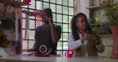 Çiçekçi telefon alırken iş arkadaşı buket hazırlıyor, kırsal kesimdeki çiçekçi dükkanında takım çalışması yapıyor, müşteri hizmetlerini dengeliyor ve çiçekçilik yapıyor.