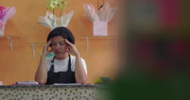 Çiçekçinin başındaki ellerle stresli görünen kadın çiçek buketleri, çiçek süslemeleri ve parlak çalışma alanındaki hediye eşyalarıyla çevrili.