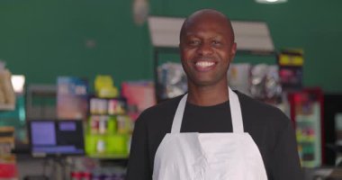 Süpermarkette önlük takan gülümseyen Afrikalı Amerikalı bir adam, renkli bir perakende ortamda doğal ışık ile neşeli, kendinden emin bir portre.