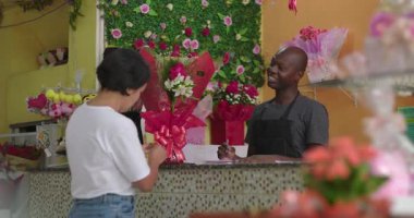 Çiçekçi tezgahında kart okuyucu ile ödeme yapan kadın müşteri. Çiçek buketleri, çiçek hediyeleri ve canlı çiçek aranjmanları ile dolu.