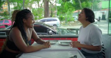 Çeşitli bayan arkadaşlar restoran masasında gülüşlerini paylaşıyorlar, neşeli samimi anlar, kapalı alanlarda günlük atmosfer şehir manzarası ve parlak doğal ışık.