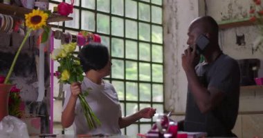 Çiçekçi telefon başında, iş arkadaşı kır çiçekçisinin içinde çiçekçilik düzenlerken iletişim ve müşteri hizmetleri hazırlıklarını dengeliyor.