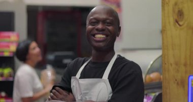 Önlüklü gülümseyen Afrikalı Amerikalı şarküteri işçisi markette kollarını kavuşturmuş dikilirken arka planda Latin kadın müşteri alışverişi yapıyor.