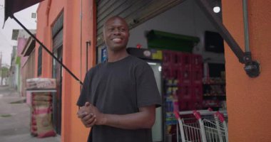 Mutlu Afrikalı Amerikalı dükkan sahibi açık kapılı mahalle marketinin önünde günün başında turuncu duvarın önünde durarak gülümsüyor.