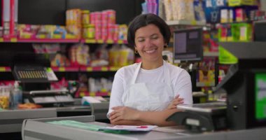 Gülümseyen kadın kasiyer, arka planda renkli raflar dolusu abur cubur, yiyecek ve market ürünleriyle dolu bir marketin kasasında güvenle oturuyor.