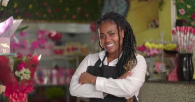 Afrikalı Amerikalı çiçekçi kollarını kavuşturuyor ve çiçekçinin içinde güvenle gülümsüyor. Renkli perakende seti içinde gururlu bir girişimci.