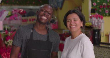 Çiçekçinin içinde mutlu bir şekilde gülümseyen çiçekçi ve müşteri, birlikte poz veren neşeli adam ve kadın çiçek aranjmanları ve renkli hediye süslemeleriyle çevrili.