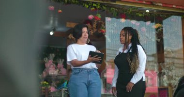 İş ortakları fikir alışverişinde bulunurken ve gülümserken çiçekçi dükkanının dışındaki dijital içeriği incelerken çiçeklerle ve dekorlarla dolu