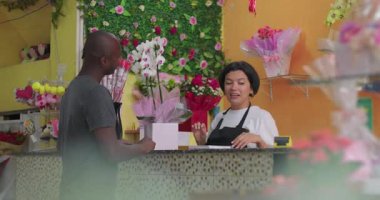 Çiçekçi dükkanında çiçekçi ve dekoratif düzenlemelerle çevrili orkide buketi için temassız kart ödeyen bir müşteri.