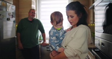 Anne kucağında bebek, baba arka planda çalışırken aile dinamiklerini ve günlük rutinlerini gözler önüne seriyor.