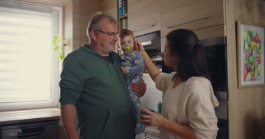 Anne, modern mutfakta babayla konuşurken bebeğin kafasına şefkatle dokunuyor. Günlük ev ortamında sıcak aile etkileşimi.