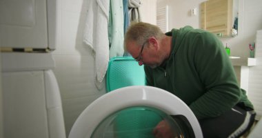 Çamaşır makinesine çamaşır sepeti ve temiz ve modern banyo ortamında havluları koymaya hazırlanırken gülümseyen bir adam.