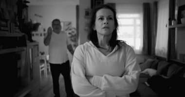 Ateşli tartışmalar sırasında kolları çapraz duran kadın kararlı ve kızgın görünürken, arka plandaki adam hayal kırıklığı içinde hareketler yapıyor, yoğun ilişki çatışmalarını yakalıyor.