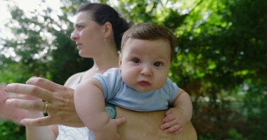 Yeşil bir parkta annesi tarafından tutulan bebek, şaşkın ve meraklı bir ifadeyle, doğal bir çevrede keşif ve keşif anını yakalıyor.