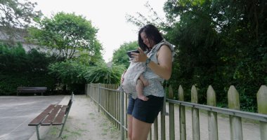 Genç anne huzurlu bir parkta akıllı telefonunu kontrol ediyor. Yavrusu rahat bir şekilde bir taşıyıcıda dinleniyor. Teknoloji ve açık havada geçirdiği güzel zamanları birleştiriyor.