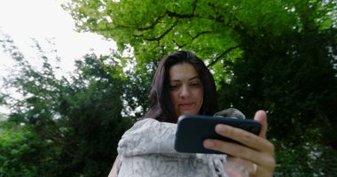 Genç anne huzurlu bir parkta akıllı telefonunu kontrol ediyor. Yavrusu rahat bir şekilde bir taşıyıcıda dinleniyor. Teknoloji ve açık havada geçirdiği güzel zamanları birleştiriyor.