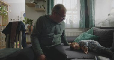 Babam, bir bebeğin yanında, emzik ile bir kanepede dinleniyor. Huzurlu bir aile salonunda, içi doğal ışıkla dolu bir el uzatıyordu.