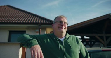 Evin önündeki konut kapısına yaslanan adam, gülümseyen ve rahat, banliyö yaşam tarzı ve mülk çevresi odaklı