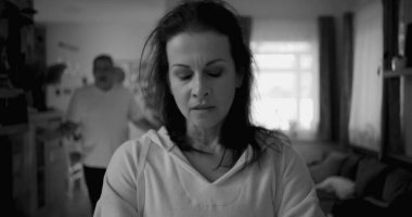 Kadın hararetli tartışmalar sırasında erkeklerden yüz çeviriyor, mesafeli ve üzgün görünüyor, erkek ise arka planda hayal kırıklığıyla işaret ediyor, duygusal gerilimi ve çatışmayı yakalıyor.