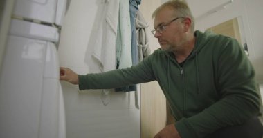 Çamaşır makinesinin önünde diz çökmüş bir adam, temiz havluları ve parlak döşeli duvarları olan modern bir banyoda çamaşırları dikkatlice yüklüyor.