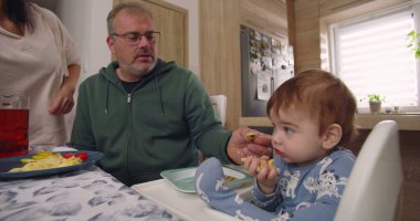 Baba yemek sırasında bebek maşasında oturur, anne tabağın yanında durur, aile bağlarını yemek masasında kurar, çocuk yemek yer.