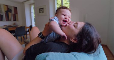 Anne sıcak ve sevgi dolu bir anda bebeği öpüyor ve kucaklıyor, doğal ışık sıcak bir oturma odasına akıyor, anne ve çocuk arasında neşeli ve samimi bir bağ kuruyor..