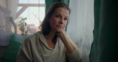 Çenesi elinde dinlenen bir kadın, derin bir düşünceyle pencereden dışarı bakıyor, yumuşak perdelerle çevrili ve evdeki sıcak elementlerle çevrili.