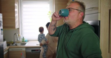 Annesi buzdolabını kollarında bebekle kontrol ederken elinde bardak tutan bir adam, tipik bir sabah rutini sırasında sıradan bir aile anı, sıcak ve besleyici ebeveynlik sahnesi.