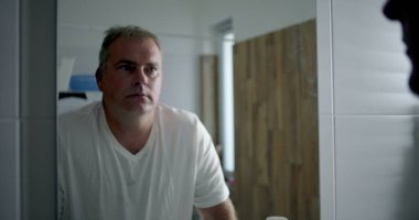 Profil görünümlü bir adam banyoda öne doğru eğiliyor, derin düşüncelere dalmış, içi beyaz fayanslarla dolu bir banyo ve ahşap kaplama, sessiz ve yansıtıcı kişisel bir an.