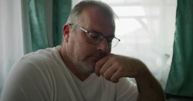 Adamın biri, çenesini elinde dinlendirirken, sakin ve rahat bir evde yumuşak yeşil perdeleri olan bir pencerenin yanında otururken düşünceli bir şekilde bakıyor.