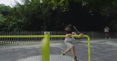 Genç bir çocuk oyun parkında enerjik bir şekilde koşuyor. Atlıkarınca, sıcak bir günde dışarıda oyun oynuyor. Etrafı yemyeşil ve park ekipmanlarıyla çevrili.