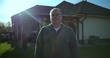 Evin arka bahçesinde duran adam, sakin bir ifade, banliyö manzarası, arka planda yeşil çimenler ve veranda, sakin atmosfer, rahat ve rahat bir an.