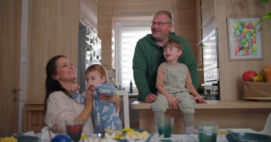 Büyük çocuk mutfak tezgahında otururken heyecanlı bir şekilde bir şeyi işaret ediyor, baba ve anne kucağında bebek, sıcak mutfakta neşeli bir aile etkileşimi.