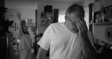 Tartışma sırasında yoğun ifadeli erkek portresi, oturma odasında arka planda kadın, duygusal gerilim ve gergin ilişki dinamiklerine odaklan, siyah-beyaz