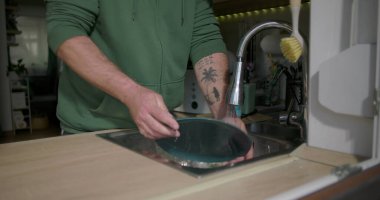 Bir adam mutfak lavabosundaki musluk suyunun altında mavi bir tabağı duruluyor, günlük ev işleri sırasında bulaşık takımlarını temizlemeye odaklanıyor.