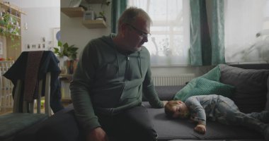 Babam, bir bebeğin yanında, emzik ile bir kanepede dinleniyor. Huzurlu bir aile salonunda, içi doğal ışıkla dolu bir el uzatıyordu.