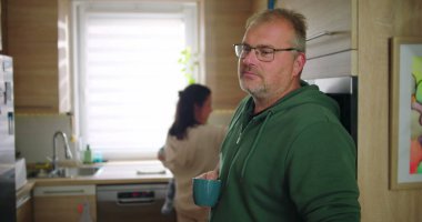 Annesi buzdolabını kollarında bebekle kontrol ederken elinde bardak tutan bir adam, tipik bir sabah rutini sırasında sıradan bir aile anı, sıcak ve besleyici ebeveynlik sahnesi.