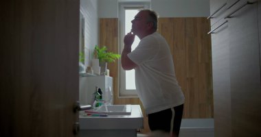 Genç çocuk girerken adam banyoda tıraş oluyor, birden fazla iş yapan ebeveyn tımara odaklanıyor, yüzüne köpük sürüyor, ahşap banyo ayrıntıları, aile sabahı rutini.