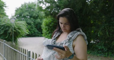 Taşıyıcıda bebeği olan bir anne telefonunun içeriğine hafif bir şok ile tepki veriyor, çoklu görev ve modern ebeveynliğin ortasında şaşırtıcı bir anın altını çiziyor.