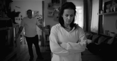 Tartışma sırasında kollarını çapraz uzatmış bir kadın, arka planda yürüyen ve el kol hareketi yapan bir adam, gergin ve duygusal ilişki çatışması, siyah ve beyaz