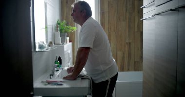 Profil görünümlü bir adam banyoda öne doğru eğiliyor, derin düşüncelere dalmış, içi beyaz fayanslarla dolu bir banyo ve ahşap kaplama, sessiz ve yansıtıcı kişisel bir an.