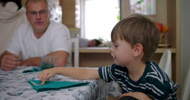 Erkek ve çocuk masada sakin bir anı paylaşırken, bir kadın arka planda çalışıyor, samimi bir evde samimi bir aile etkileşimi sergiliyor.