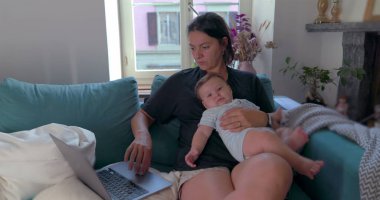 Anne kucağında bebeği kucağında tutarken rahat bir oturma odasında dizüstü bilgisayarla çalışıyor, işini ve aile zamanını aynı anda yönetiyor, görevlerine odaklanıyor.