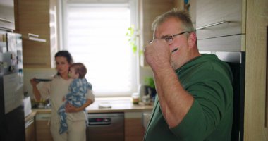 Anne bebeğini arka planda tutarken, adam mutfakta içki karıştırıyor. Aile sabah rutini sırasında birden fazla iş yapıyor. Günlük ev hayatı.