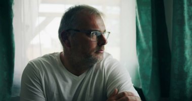 Pencere kenarında oturan bir adam, düşünceli bir ifadeyle dışarı bakıyor, kollarını kavuşturmuş, rahat bir duruş içinde, sakin bir iç mekanda yumuşak perdelerle çevrili.