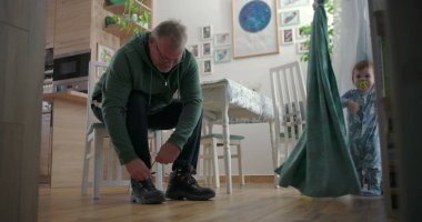 Babam, sıcak ahşap döşemeleri ve doğal ışıklandırması olan sıcak bir aile yemek odasında, emzik ile perdelerin yanında duran bir çocuğa bakarken botlarını bağlıyor.