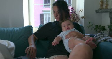 Anne evde dizüstü bilgisayarla çalışırken bebeğini kucağında tutuyor, çocuk bakımını dengeliyor ve rahat bir oturma odasında uzaktan çalışıyor.