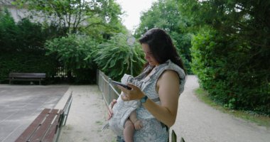 Anne, bebeğiyle birlikte parkta duruyor. Telefonuna dalmış vaziyette. Teknoloji ve ebeveynliğin günlük aktivitelerdeki karışımını örnek alıyor.